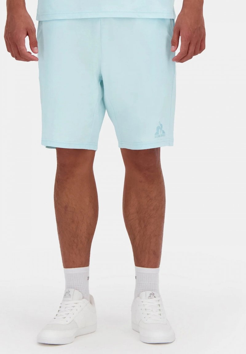 Pantalones cortos de color azul claro, hechos de una tela suave, con un corte relajado y un logotipo bordado en la parte inferior izquierda. Combinados con zapatos blancos.