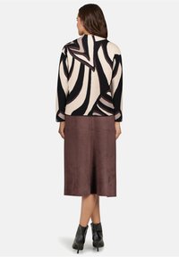 Vrouw staand met de rug naar de camera, draagt een zwarte en crèmekleurige trui met abstract patroon, een bruine midi-rok en zwarte enkellaarzen.