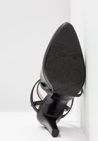 Chaussure à talons hauts noire avec un bout pointu et des sangles texturées. La semelle présente un motif en relief avec des pièces de monnaie et des motifs complexes.