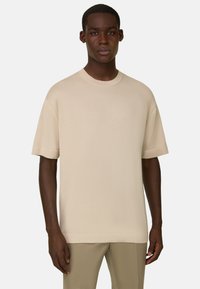 Homme à la peau foncée portant un pull  à manches courtes beige uni et un pantalon marron clair, debout devant un fond gris clair uni.