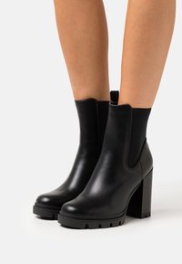 Botas de tobillo de cuero negro con un tacón grueso en bloque, paneles laterales elásticos, punta redonda y suela de goma texturizada. Acabado suave en toda la superficie.