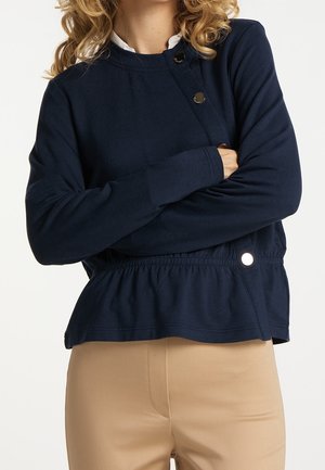Kobieta w granatowym długim rękawie z zatrzaskami i falbaną na dole, ubrana w beżowe spodnie, ze skrzyżowanymi ramionami.