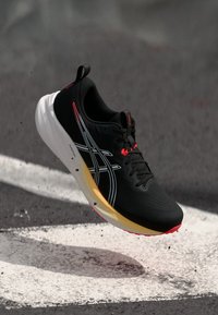 Svart sporty sko med hvite og røde detaljer, teksturert overdel, strømlinjeformet design, og en multikolor såle som går fra gult til rødt.