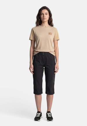Femme portant une chemise beige à manches courtes et un pantalon noir aux genoux, debout sur un fond blanc uni.