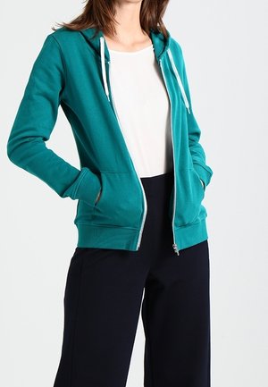 Femme portant un sweat à capuche zippé bleu sarcelle avec des cordons blancs, par-dessus une chemise blanche et un pantalon noir, mains dans les poches du sweat.