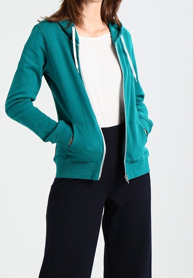 Femme portant un sweat à capuche zippé bleu sarcelle avec des cordons blancs, par-dessus une chemise blanche et un pantalon noir, mains dans les poches du sweat.