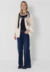 Crème overhemd met knoopjes over een zwarte top, gecombineerd met donkerblauwe wijduitlopende jeans. Accessoires zijn een gestreepte zwart-beige sjaal. Zwarte laarzen.