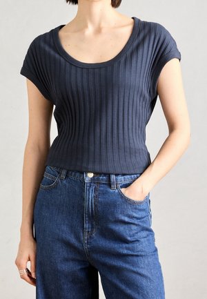 Frau trägt ein marineblaues, geripptes Kurzarmoberteil, das in hoch taillierte blaue Jeans aus Denim gesteckt ist, mit einer Hand in der Tasche und silbernen Ringen an der anderen.