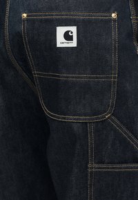 Σκούρο μπλε ύφασμα τζιν με τετράγωνη τσέπη που διαθέτει κίτρινη ραφή, χάλκινα κόλλητρα και λευκή ετικέτα λογότυπου Carhartt.