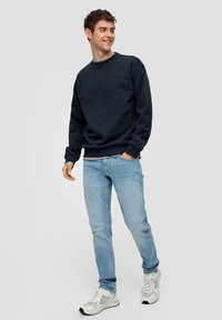 Felpa blu navy con polsini e orlo a coste, abbinata a jeans slim-fit azzurri e sneakers bianche con dettagli grigi.