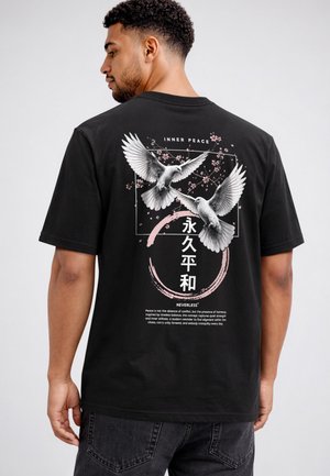 Mann trägt ein schwarzes T-Shirt mit einer Grafik von zwei weißen Tauben, pinken Blumen und Text inklusive "Inner Peace" und asiatischen Zeichen auf dem Rücken.