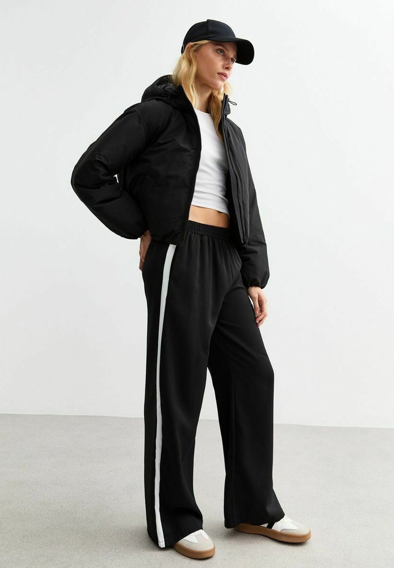 Chaqueta acolchada negra con capucha, top corto blanco y pantalones de chándal negros con rayas laterales blancas. Calzado casual completa el look.