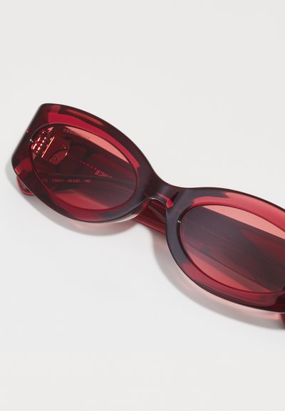 Red semi-transparent sunglasses with oval lenses, thick frames, and subtle branding on the inner temple. Smooth finish and stylish curves.