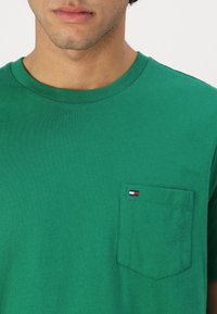 Zelené bavlněné tričko s kulatým výstřihem, krátkými rukávy a náprsní kapsou se zdobením malého loga v červené, bílé a modré barvě.