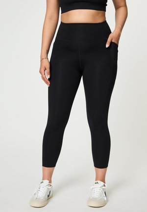 Naine kannab musti kõrge vöökohaga retuuse küljetaskuga, musta lühikest topi ja valgeid tenniseid helel ühtlasel taustal.