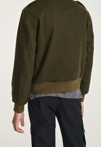 Felpa verde oliva con scollo rotondo, polsini e orlo a costine, indossata sopra una maglietta grigia, mostrata di spalle.