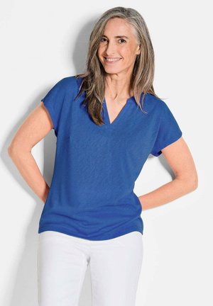 Blaues V-Ausschnitt-Bluse aus leichtem Material mit kurzen Ärmeln, featuring einer Tasche auf der linken Seite und einem lockeren Schnitt. Getragen mit weißen Hosen.