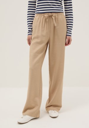 Persoon draagt beige broek met wijde pijpen en een taille met trekkoord, witte sneakers en een navyblauw-wit gestreept shirt met lange mouwen.