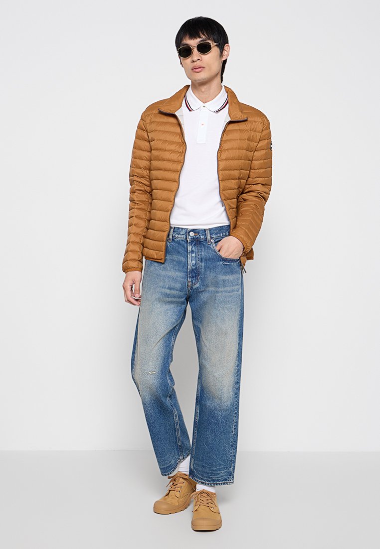 Uomo che indossa una giacca a vento marrone, una polo bianca, jeans blu larghi, scarpe marroni e occhiali da sole scuri, in piedi contro uno sfondo bianco.