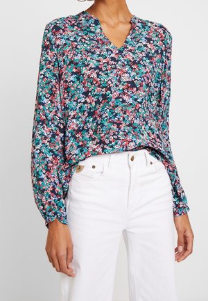 Femme portant un chemisier à fleurs ample à manches longues avec un col en V, rentré dans un pantalon blanc taille haute, debout devant un fond uni.