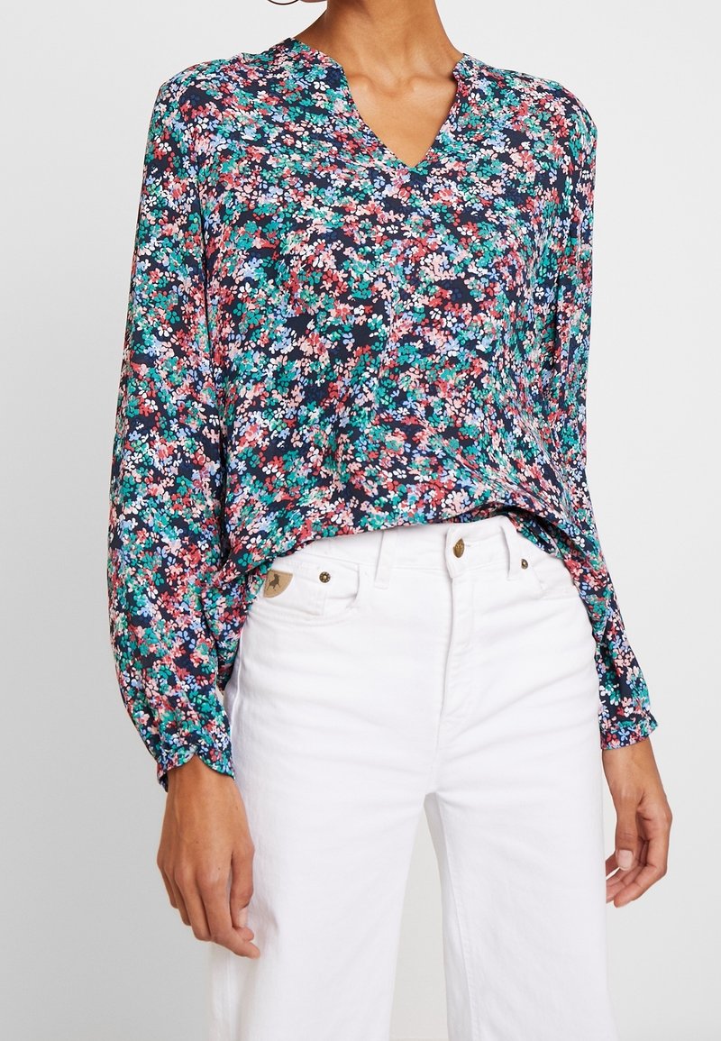 Femme portant un chemisier à fleurs ample à manches longues avec un col en V, rentré dans un pantalon blanc taille haute, debout devant un fond uni.