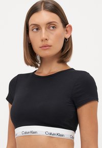 Svart cropped t-skjorte laget av glatt stoff. Har korte ermer og en hvit elastisk kant med "Calvin Klein" merkevare ved kanten.