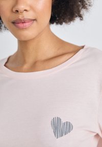 Ljust rosa t-shirt i mjukt tyg, med en grårandig hjärt-grafik på vänster bröstområde och en bred halsringning.