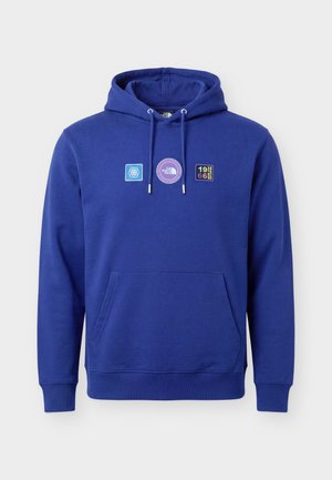 Mand iført en blå hættetrøje med frontlomme og tre små patches på brystet, stående mod en ensfarvet lys baggrund.