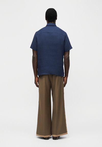 Άνδρας που στέκεται με την πλάτη προς το μέρος μας, φορώντας μπλε ναυτικό πουκάμισο με κοντά μανίκια και καφέ παντελόνι με φαρδιά πόδια, σε απλό λευκό φόντο.