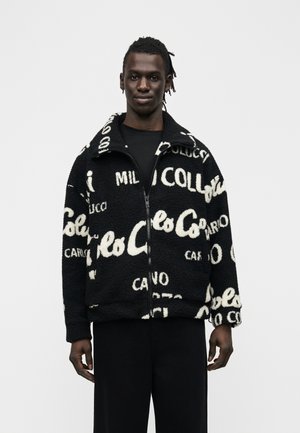 Młody mężczyzna o ciemnej karnacji, ubrany w czarno-białą polarową kurtkę na zamek z wzorem oraz czarne spodnie, stoi na jednolitym tle.