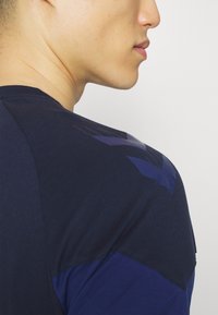 Mörkblå T-shirt med en mjuk textur, korta ärmar och ett subtilt geometriskt mönster på axlarna. Närbild från sidan.