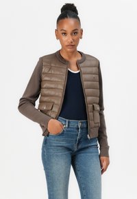 Braune zip-up Jacke mit gestepptem Vorderteil, kontrastierenden Ärmeln und geripptem Kragen. Über einem dunklen Oberteil getragen und mit blauen Jeans kombiniert.
