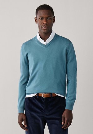 Hombre con un suéter azul claro de cuello en V sobre una camisa blanca, pantalones oscuros y un cinturón marrón, de pie frente a un fondo liso y claro.