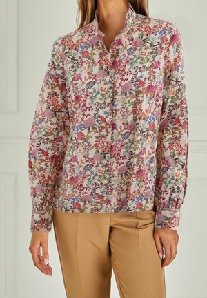 Woman wearing a long-sleeve button-up blouse with a multicolored floral pattern and beige pants, standing indoors against a white wall.