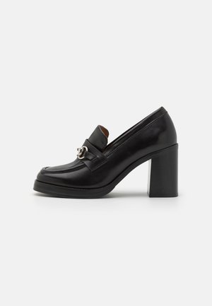 Mocasines de tacón alto de cuero negro con punta redonda, detalle de herrajes de plata y tacón cuadrado grueso. Textura suave y diseño elegante.