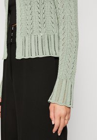 Manche de cardigan tricoté vert clair avec motif de feuille portée avec un pantalon noir taille haute, main détendue sur le côté avec une bague en or au doigt.