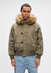 Hombre vistiendo una chaqueta bomber verde oliva con capucha forrada de piel y puños acanalados, combinada con jeans de color azul claro frente a un fondo liso.
