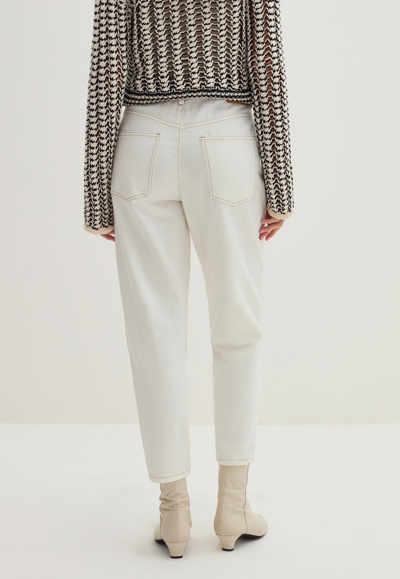 Rückansicht einer Person, die weiße High-Waist-Hosen, beigefarbene Stiefeletten mit niedrigen Absätzen und einen schwarz-weißen gemusterten Strickpullover trägt.