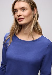 Bluzka z długim rękawem w jednolitym niebieskim kolorze o gładkiej fakturze i zaokrąglonym dekoltem. Charakteryzuje się minimalistycznym designem, bez wzorów i ozdób.