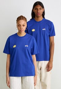 Twee individuen dragen levendige, blauwe katoenen t-shirts met grafische ontwerpen: de ene met een geel exploderend patroon, de andere met een klein logo.