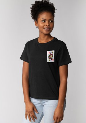 Junge Frau mit lockigem Haar, die ein schwarzes T-Shirt mit einer Spielkarte Dame Herze auf der Brust trägt.