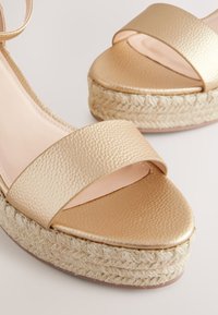 Sandales compensées dorées avec des brides en cuir lisse et une semelle en jute tressé. La tige présente une finition texturée et un large bout ouvert.