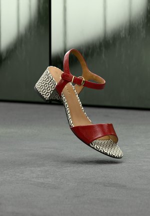 Red leather sandal with a block heel, featuring a patterned black and white sole and a buckle strap. Smooth texture and open toe design.