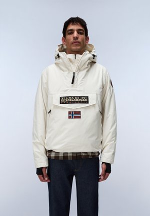 Mand iført cremefarvet Napapijri hættetrøje med frontlomme og norsk flagpatch, mørke jeans og ternet skjorte under.