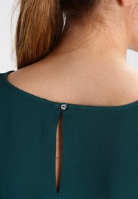 Teal blouse van textiel met structuur, voorzien van een keyhole uitsparing aan de achterkant, vastgezet met een klein knoopje bij de hals.