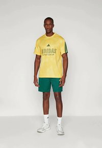 Rumena majica Adidas z zelenim napisom, akcenti in črtami, v kombinaciji z zelenimi kratkimi hlačami. Obutev so bele športne čevlje.