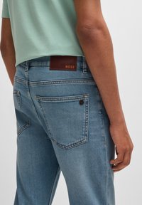 Calças de ganga azul claro com uma etiqueta de marca em couro castanho e dois bolsos traseiros. As calças têm um corte reto e costuras contrastantes.