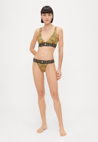 Lāču raksta bikini komplekts, kas ietver trīsstūru krūšturi un saderīgus apakšbikses, akcentētas ar melnu joslu un zelta logo detaļu.