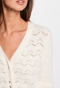 Cardigan bianco lavorato a maglia con scollatura a V, caratterizzato da un design a zigzag con ritagli, texture morbida e piccoli bottoni metallici.