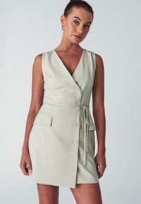 Vestido sin mangas de color verde claro con escote en V, diseño envolvente, bolsillos laterales, botones y una cinta en la cintura, hecho de una tela suave y texturizada.